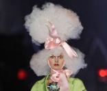 Gros plan sur une catwalk du défilé printemps-été 2009 de John Galliano. Le modèle perché sur de très hauts talons et coiffé d'une incroyable perruque blanche,  porte une veste de couleur acidulée sur une petite jupe fleurie. 