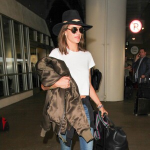 Alessandra Ambrosio à l'aéroport LAX à Los Angeles, porte un chapeau ále by alessandra, un t-shirt court blanc, un jean RE/DONE et des bottines Saint Laurent. La Brésilienne tient à la main un bomber Ben Taverniti Unravel Project. Le 8 février 2016.