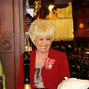 Barbara Windsor (en tailleur rouge) recevant la visite de la reine Elizabeth II et du duc d'Edimbourg sur le tournage du soap opera EastEnders en novembre 2001 à Londres.