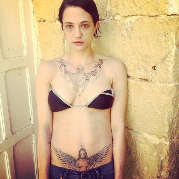 Asia Argento avec deux de ses plus gros tatouage, son collier victorien en haut de la poitrine, et son ange au bas du ventre (2014).