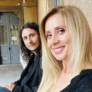 Assis sur un banc, le couple est très élégant : Lara Fabian porte une robe noire à dentelle alors que son époux est vêtue d'une veste de costume noir et d'un jean brut.