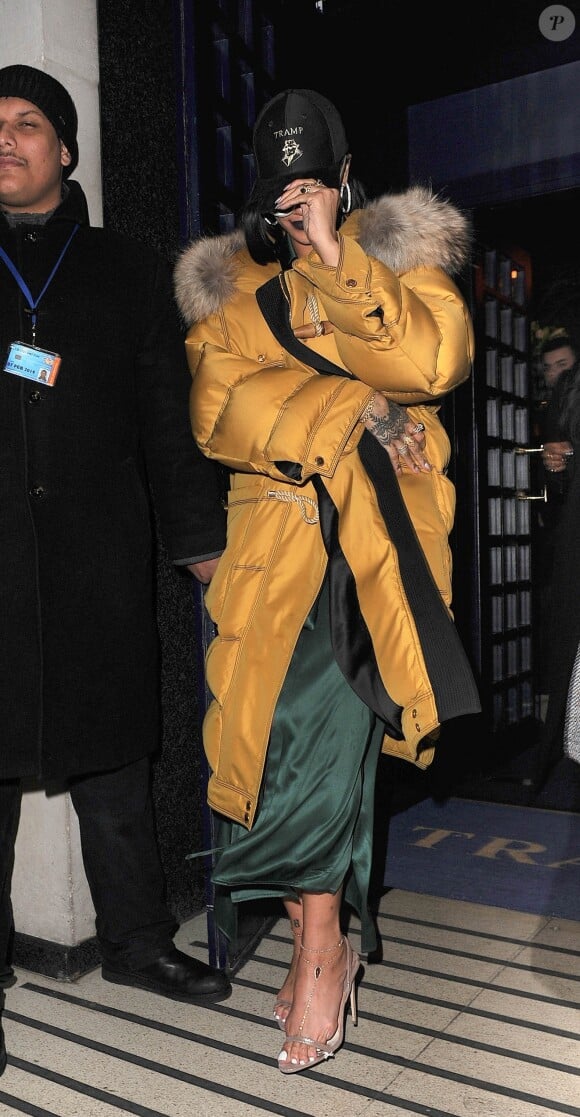 Rihanna quitte le Tramp à Londres, habillée d'un duffle-coat jaune Burberry et d'une robe en soie Rosetta Getty. Une casquette à l'effigie de la boîte de nuit londonienne Tramp et des lunettes de soleil Adam Selman x Le Specs accessoirisent sa tenue. Le 24 février 2016.