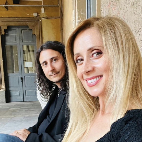 Assis sur un banc, le couple est très élégant : Lara Fabian porte une robe noire à dentelle alors que son époux est vêtue d'une veste de costume noir et d'un jean brut.