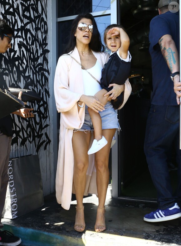 Kourtney Kardashian porte sa fille Penelope Disick à Miami, habillée d'un gilet couleur chair, d'un body blanc, d'un mini-short en jean Levi's et de sandales Stuart Weitzman. Des lunettes de soleil Victoria Beckham et un sac Givenchy (modèle Pandora Box) accessoirisent sa tenue. Le 24 avril 2016.