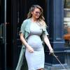 Chrissy Teigen, enceinte et en balade à New York, porte des lunettes de soleil Victoria Beckham, un trench-coat 3.1 Phillip Lim, une robe grise et des sandales. Le 16 novembre 2015.