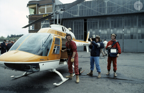 Archives - En France, Philippe de Dieuleveult, devant l'hélicoptère, vêtu de sa combinaison rouge, portant son casque d'écoute et son poste émetteur, le cameraman, Patrick FABRY et le responsable du magnétoscope, Serge BEAUPERE lors de l'émission La chasse aux trésors en mai 1983