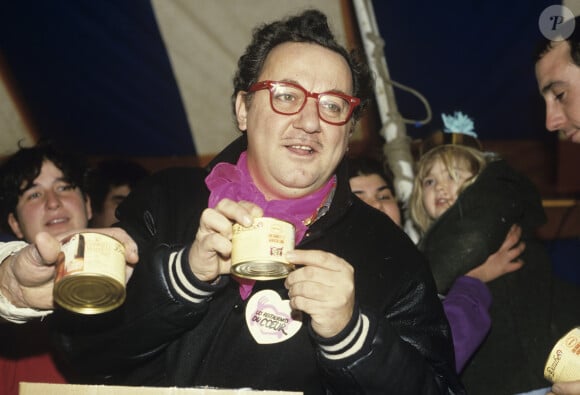 Archives - En France, à Genevilliers, sous le chapiteau des Restaurants du coeur, lors d'une distribution de nourriture, entouré de bénévoles, Coluche arborant un badge de l'association sur son blouson le 3 janvier 1986.