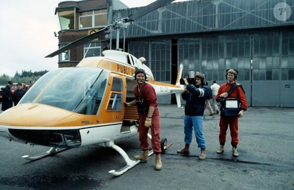 Archives - En France, Philippe DE DIEULEVEULT, devant l'hélicoptère, vêtu de sa combinaison rouge, portant son casque d'écoute et son poste émetteur, le cameraman, Patrick FABRY et le responsable du magnétoscope, Serge BEAUPERE lors de l'émission La chasse aux trésors en mai 1983