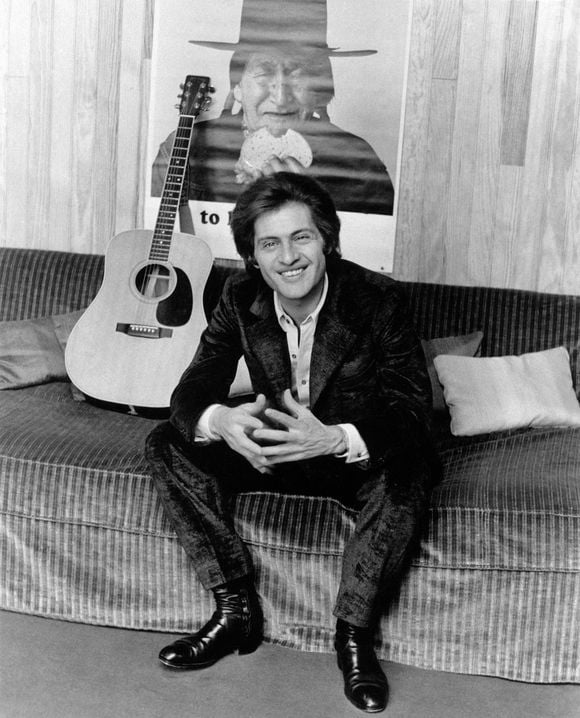 Le chanteur français d'origine américaine Joe Dassin sourit sur un canapé à côté d'une guitare. Derrière lui, une affiche représentant un Indien d'Amérique. Paris, 1970 Photo par Agenzia Pitre/Mondadori Portfolio via ZUMA Press/ABACAPRESS.COM