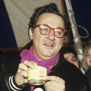 Archives - No Web No chaines TV - En France, à Genevilliers, sous le chapiteau des RESTAURANTS DU COEUR (LES RESTOS DU COEUR), lors d'une distribution de nourriture, entouré de bénévoles, Coluche arborant un badge de l'association sur son blouson le 3 janvier 1986.
