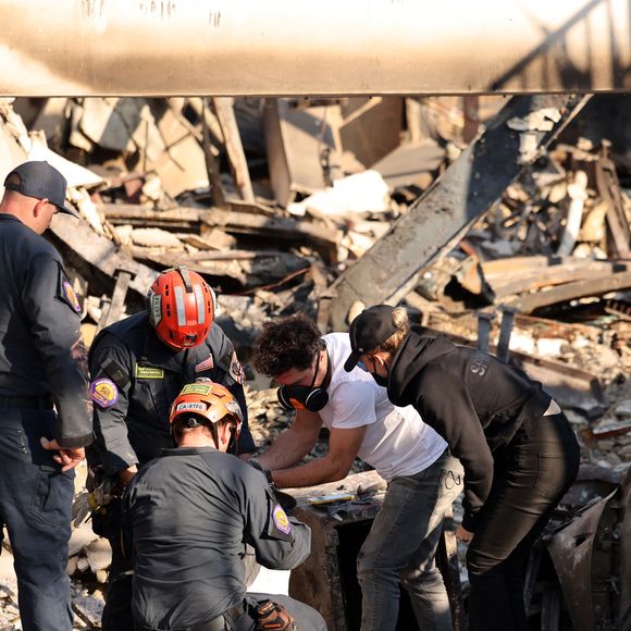 Exclusif - Prix Spécial - No Web - Des pompiers bienveillants, en déploiement dans sa rue, sont venus prêter main-forte à Laeticia Hallyday en la voyant essayer de rassembler quelques affaires - Laeticia a enfin pu pénétrer au cœur de Pacific Palisades pour constater de ses propres yeux les dégâts causés par les pires incendies de l’histoire de Los Angeles. À peine arrivée sur les ruines de sa maison, elle découvre l’ampleur du désastre : elle s’effondre en larmes en passant devant son église dévastée, puis observe tout le quartier, les maisons des voisins, et enfin la sienne, complètement ravagée par les flammes. Même les poutres porteuses, pourtant très résistantes, sont tombées ou tordues sous l’effet de la chaleur intense. Seule face à cette nouvelle épreuve, Laeticia se blottit auprès de Joy, venue la soutenir. Ensemble, elles contemplent un amas de souvenirs réduits en cendres et une montagne de braises. L’air est lourd et nocif, et la gorge gratte après seulement quelques minutes sur place. Le décor apocalyptique du quartier, autrefois vivant et animé avec ses commerces et son voisinage, pèse lourdement. Désormais, un silence absolu règne dans ce lieu où la qualité de vie était sans pareil. Des pompiers bienveillants, en déploiement dans sa rue, sont venus prêter main-forte à Laeticia en la voyant essayer de rassembler quelques affaires. Ils s’assurent que tout le monde a été épargné par l’incendie, car la zone est désormais inspectée par les autorités locales comme une zone de crime. En effet, on continue de retrouver des corps, et il reste encore plusieurs dizaines de disparus.Tout le secteur est d’ailleurs bouclé par des checkpoints tenus par la garde nationale de l’armée américaine, équipée d’uniformes de guerre, de Hummers et d’autres véhicules militaires. Sous sa casquette, son masque et ses lunettes, Laeticia pleure toutes ses larmes face à son cocon parti en fumée. Son ami l’aide à chercher d’éventuels objets récupérables et finit par retrouver son coffre qui semble avoir survécu aux flammes. Mais en le déplaçant, la porte s’ouvre soudainement, révélant que tout ce qu’il contenait a été consumé. Le peu d’espoir de Laeticia de retrouver ses objets de valeur disparaît en fumée. Tant bien que mal, elle tente de récupérer quelques bijoux calcinés et le téléphone de Johnny qu’elle conservait précieusement. Tout cela se déroule au pieds de ses deux voitures complètement brûlées garées devant la maison : la voiture de Jade et la Bentley de Laeticia. Cependant, son ancienne Audi, garée de l’autre côté de la rue, est restée intacte. Laeticia passe une bonne partie de l’après-midi à fouiller les décombres, un peu hagarde. Chaque recoin de la maison lui rappelle des souvenirs de bonheur et la fait éclater en sanglots. Elle repart avec un nœud dans l’estomac, avouant à peine dormir la nuit à cause du stress et du choc. Les incendies sont fréquents dans la région, mais Laeticia confie avoir cru qu’elle pourrait revenir dans sa maison dès le soir même après l’ordre d’évacuation. C’est pour cette raison que Joy n’avait pris que le strict minimum en emmenant leur chienne, car personne à Pacific Palisades n’imaginait un tel désastre possible. Dans le quartier de Pacific Palisades, à Los Angeles, Californie, Etats-Unis, le 18 janvier 2025.  POUR INFO VOIR VENDEUR